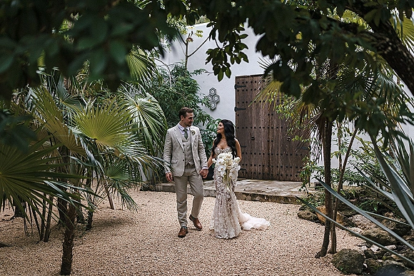 Couple portrait of bride and groom walking hand in hand, bride with cascading orchid bouquet, beside palm trees and a white stucco wall