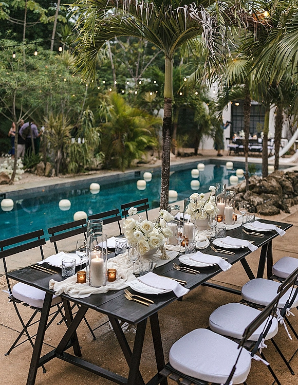 Reception tablescape set for a poolside wedding reception with white floral centerpieces, candles in hurricane vases, and string lights by palm trees