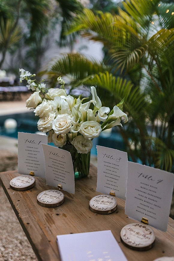 Wedding escort cards with calligraphy escort cards in gold holders, surrounded by white roses, calla lilies, and greenery on a poolside patio table