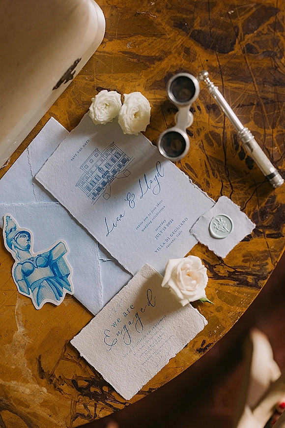Wedding invitation suite with a save the date card on a marble tabletop, deckle-edge paper, envelope with wax seal, white roses, pen and opera glasses