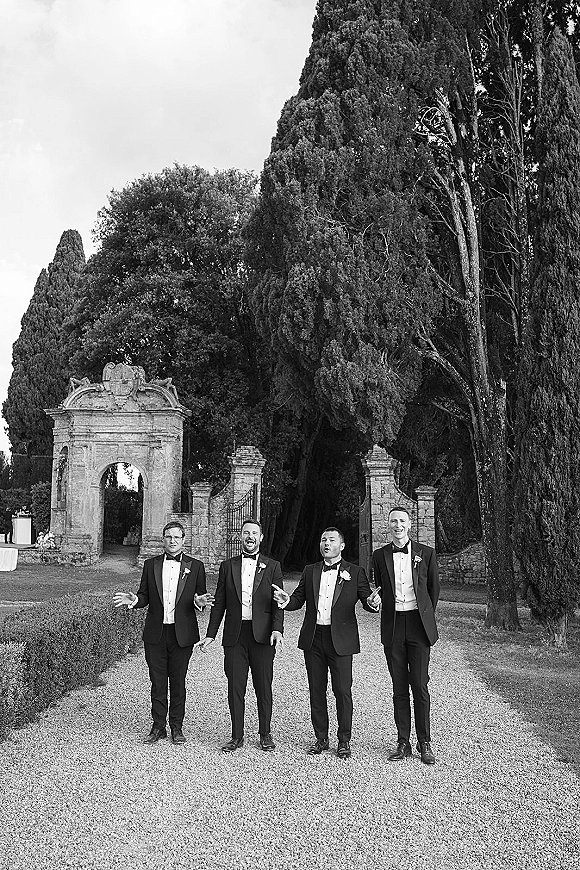 Groomsmen portrait of men in tuxedos walking and laughing in black tuxedos with boutonnieres on a gravel path by a stone archway