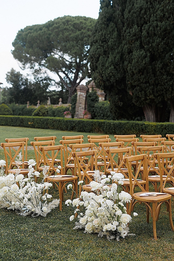 Outdoor ceremony setup with wood cross-back chairs lined in rows, white aisle florals and programs on seats on a garden lawn