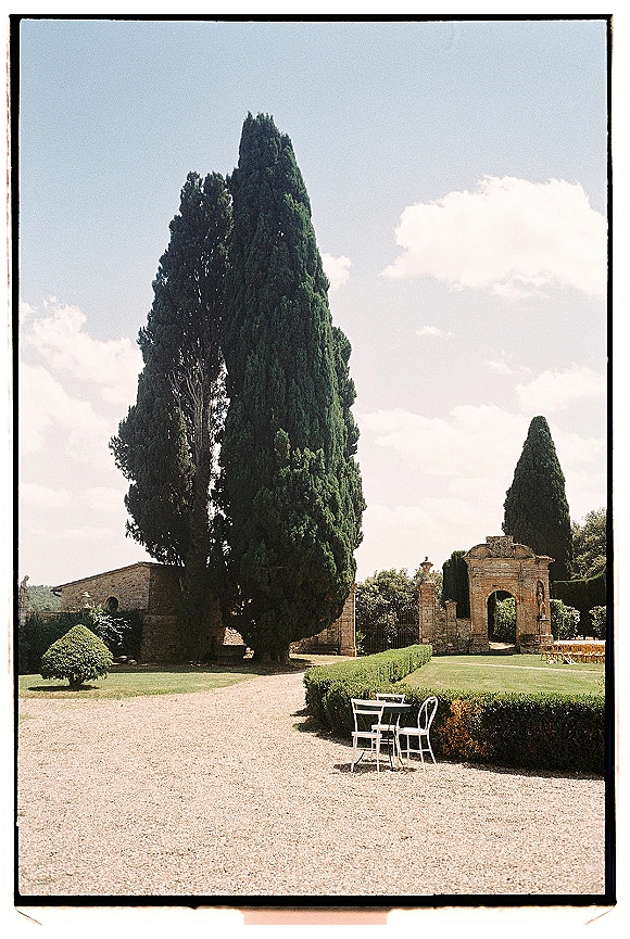 Outdoor wedding venue with white bistro chairs and table on a gravel path, framed by a stone archway and cypress-lined hedges under blue sky