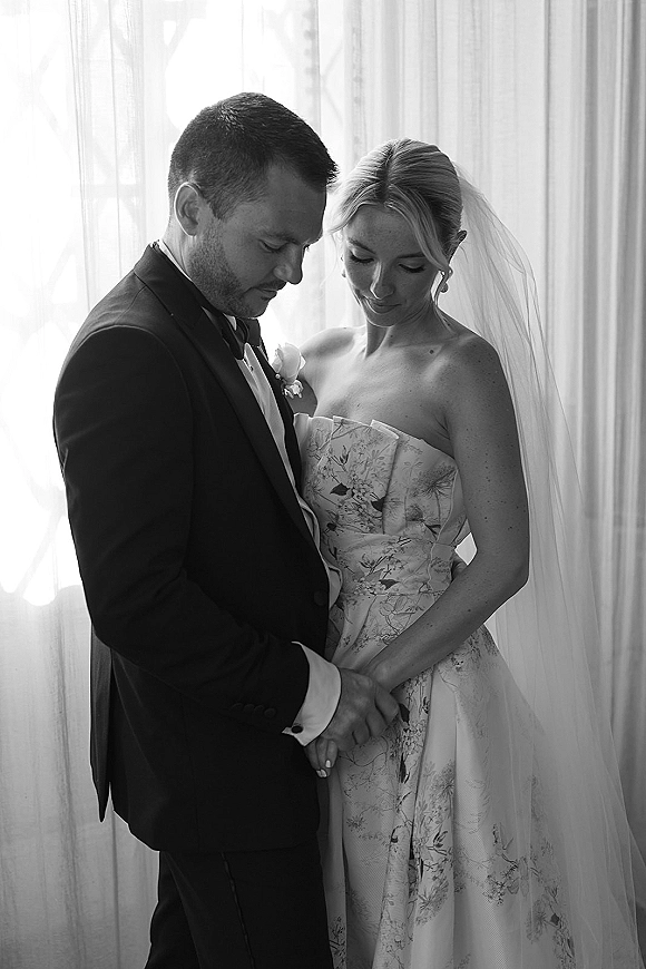 Couple portrait of bride and groom portrait holding hands by a window, her strapless dress and veil glowing in soft indoor light