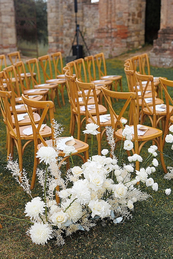 Ceremony seating with outdoor ceremony chairs in curved rows, wooden chairs lined with white florals, petals, and programs on a lawn by a stone wall