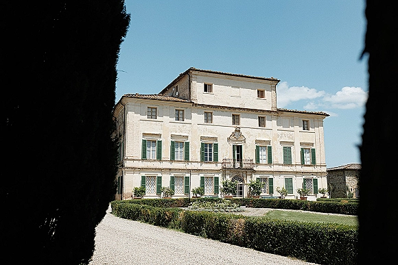 Wedding venue exterior of a villa wedding venue with balcony and green shutters, framed by potted plants and hedges on a gravel drive under blue sky