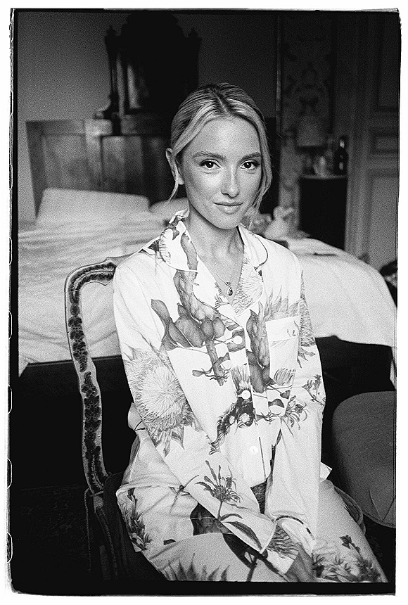 Bridal getting ready portrait of a bride in pajamas, wearing a necklace and earrings while seated in an upholstered chair in a bedroom