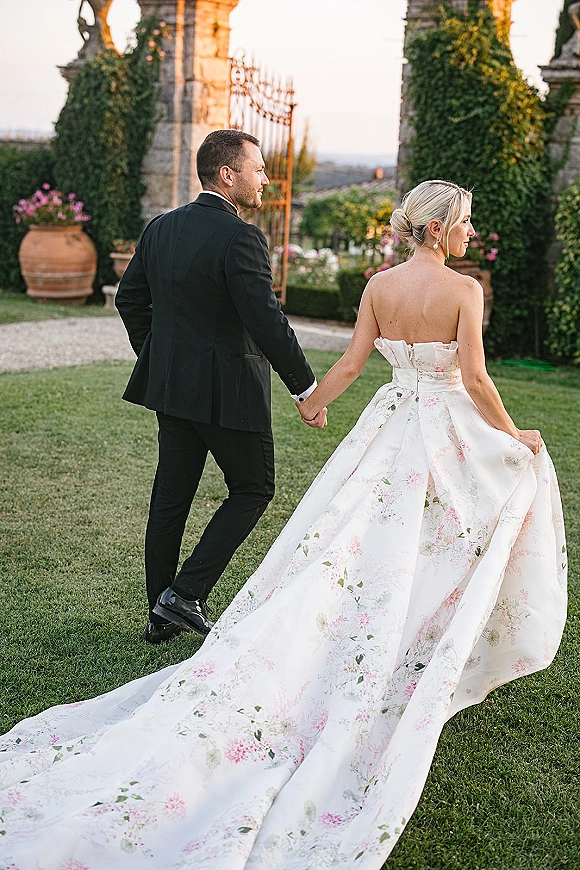Couple portrait of bride and groom holding hands, walking through a garden gate at sunset, her strapless gown train flowing behind