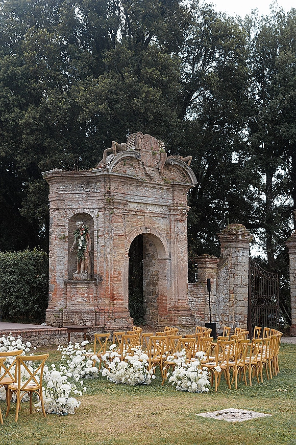 Outdoor ceremony setup with garden wedding ceremony seating of cross back chairs and white aisle flowers before a stone arch in ruins