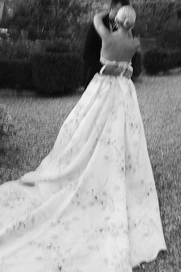 Wedding couple portrait of bride and groom hugging, her strapless dress with long train flowing on a garden lawn by hedges and path
