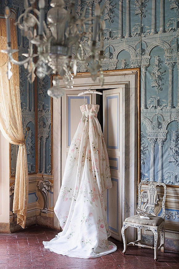 Wedding dress hanging wedding dress with floral print on a hanger beneath an ornate chandelier, shoes on a chair in a vintage room