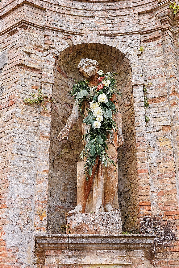 Statue floral garland of white and red roses with greenery draped in a stone arch niche against a rustic brick wall outdoors