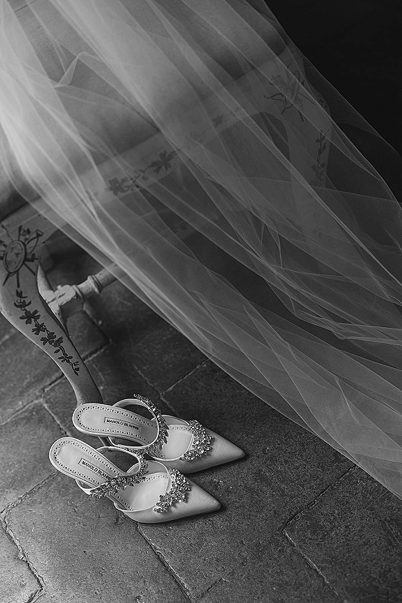 Wedding shoes with rhinestone buckle slingback heels beside a bridal veil on a stone floor near a vintage chair