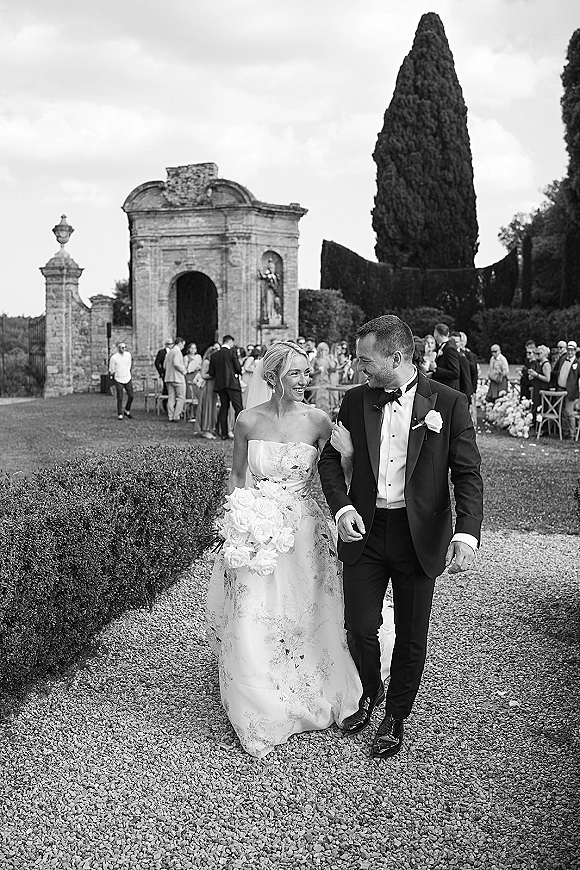 Wedding couple portrait of bride and groom walking arm in arm, bride holding a bouquet with veil, along a garden walkway by a stone archway