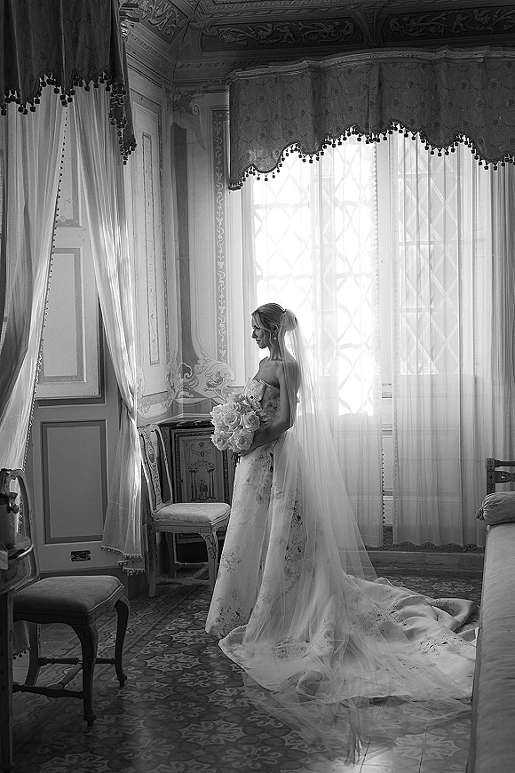 Bridal portrait in black and white of a bride by window in a strapless wedding dress with long cathedral veil, bouquet, and earrings in an ornate room