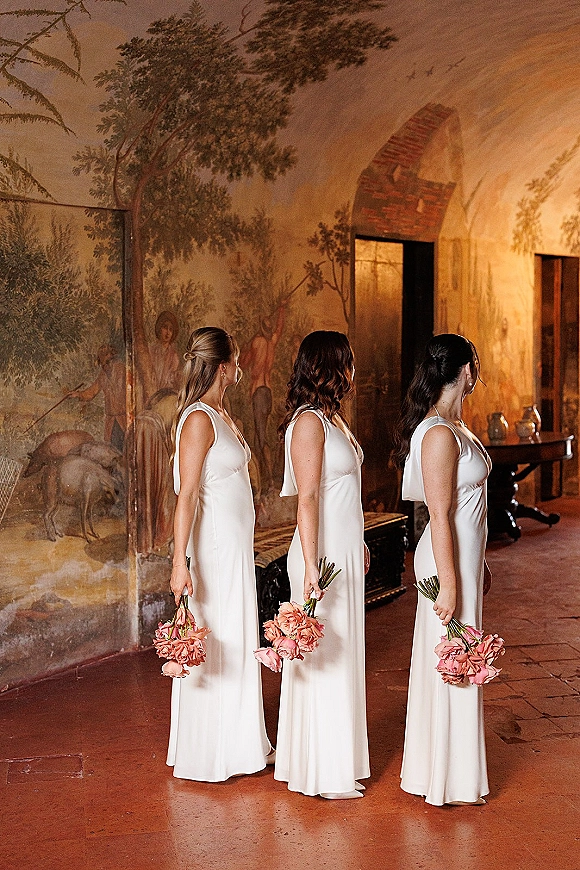 Bridesmaid portrait of three women in white satin dresses holding pink rose bouquets by a fresco mural wall and arched doorway