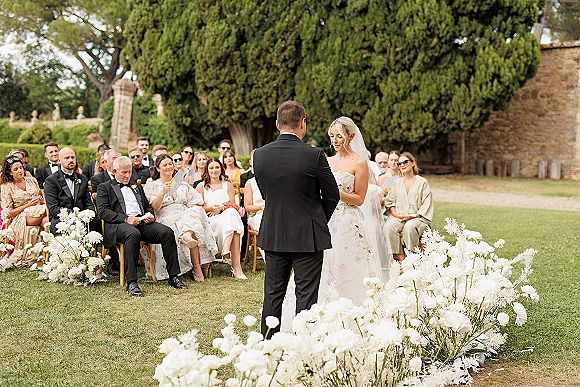 Wedding vows during an outdoor wedding ceremony as bride in veil and strapless dress reads to groom in tux, guests seated on garden lawn