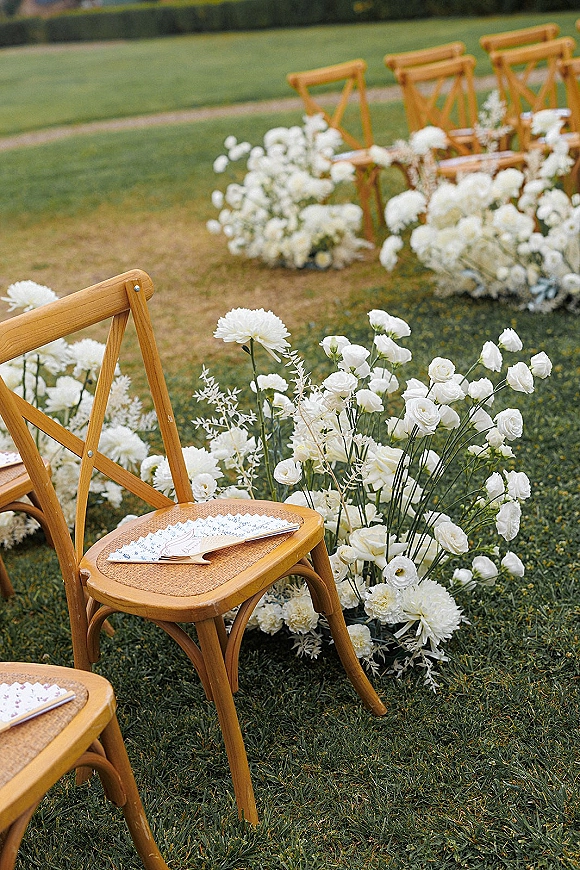 Ceremony aisle decor with outdoor ceremony aisle flowers lining a grassy pathway, cross back chairs with programs and white floral clusters near a hedge