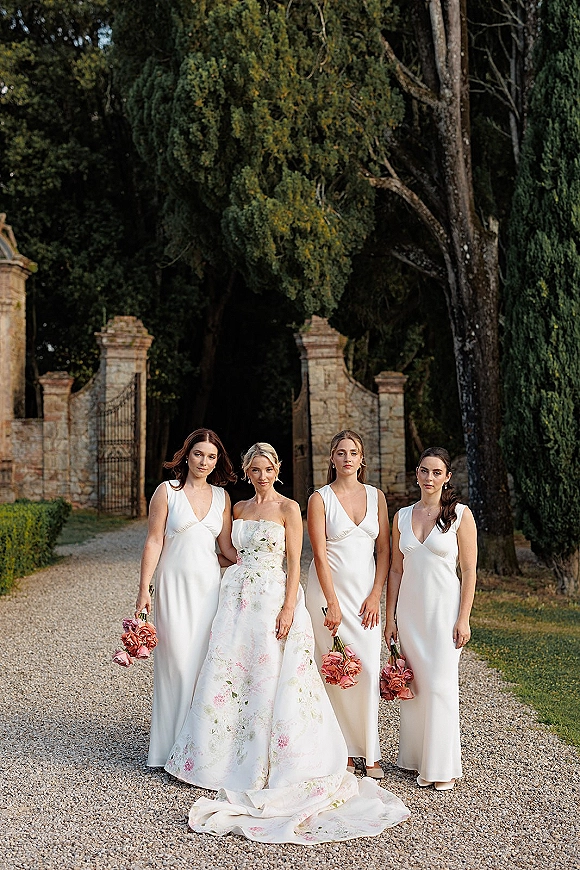 Bride and bridesmaids in a bridal party portrait walking with pink bouquets along a gravel path by a stone gate and hedges