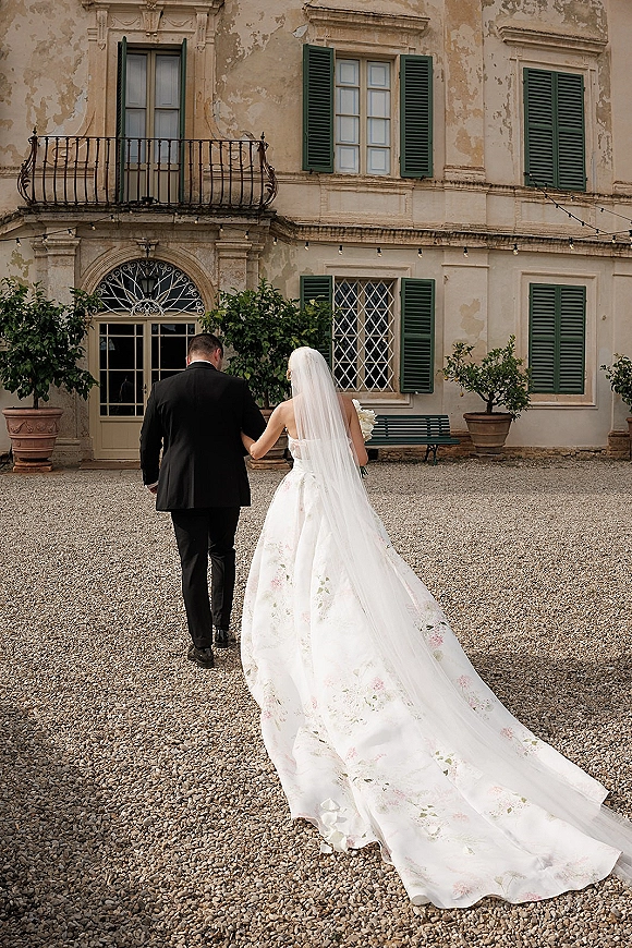 Wedding couple walking away under courtyard string lights, bride in embroidered gown with cathedral veil holding bouquet by villa facade