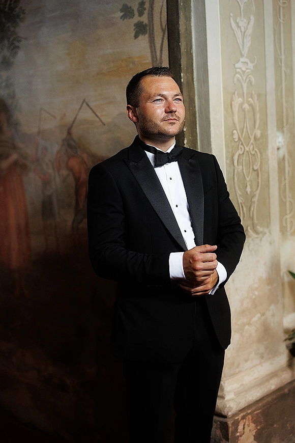 Groom portrait in a classic black tuxedo and bow tie, hands clasped, looking away beside an ornate wall mural and doorway indoors