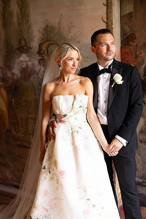 Couple portrait of bride in strapless floral wedding dress and veil with groom in black tuxedo, posing by a painted mural wall indoors