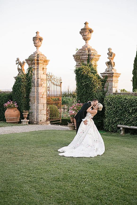 Wedding kiss portrait of bride and groom kissing in a dip, her dress train flowing by an ivy-covered wrought iron gate with statues behind