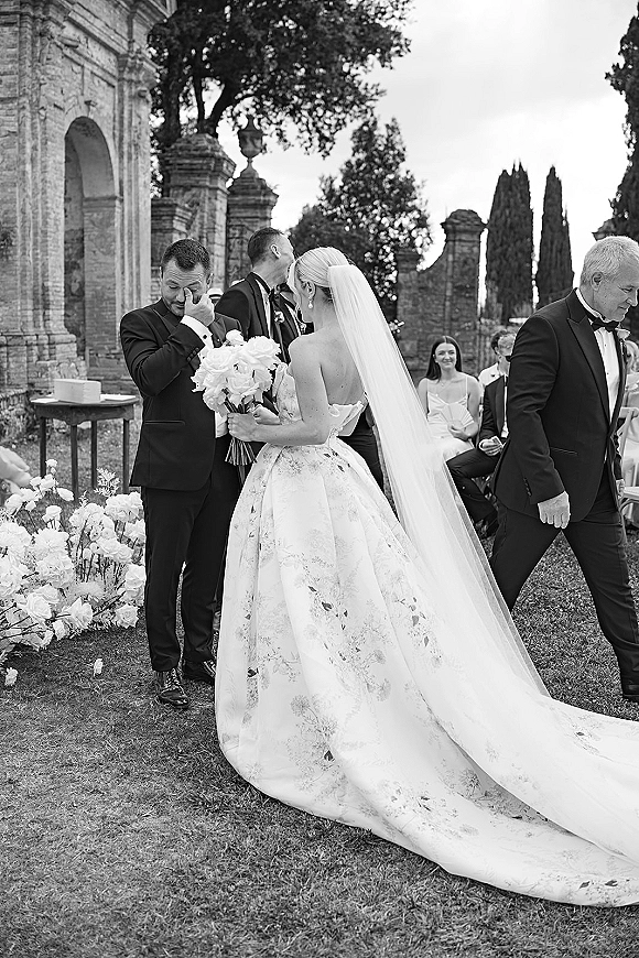 Ceremony moment as groom wiping tears while facing bride from behind in a cathedral veil, with guests by aisle flowers at stone pillars