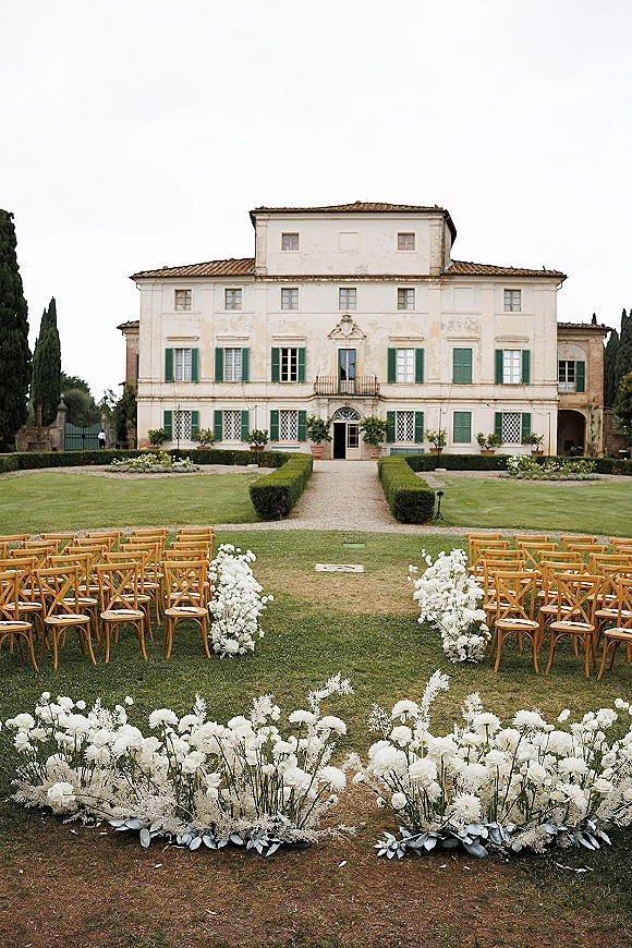 Outdoor ceremony setup with wood crossback chairs lining a gravel aisle, white floral arrangements and greenery before a villa facade