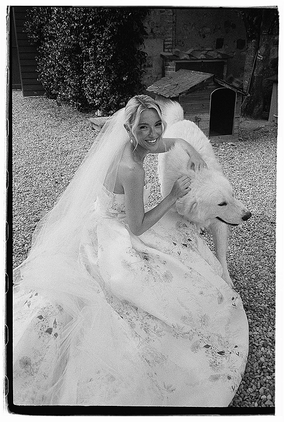 Bridal portrait of a bride with dog, sitting on gravel in a strapless embroidered gown and veil beside a wooden doghouse and shrubs
