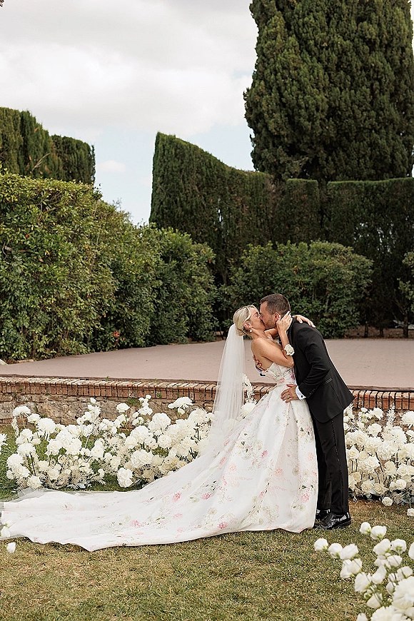 Wedding kiss portrait of bride and groom kissing as her veil and floral train spread, framed by garden hedges and a brick wall under cloudy sky