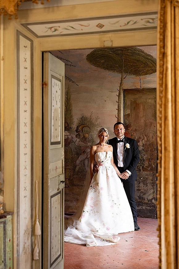 Couple portrait of bride in strapless floral gown with long veil beside groom in black tuxedo, posed by a painted mural wall doorway indoors