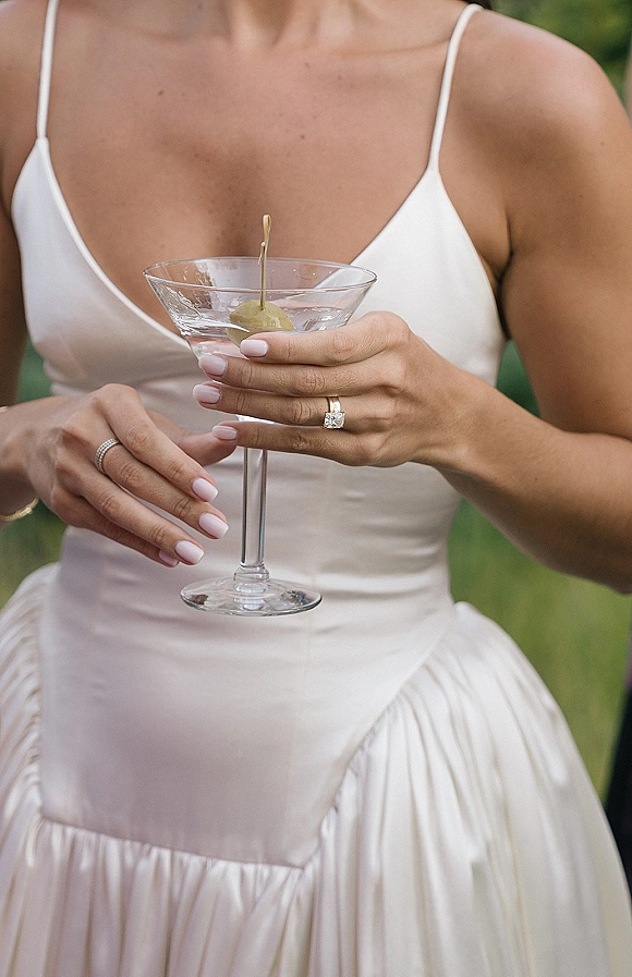Bridal cocktail detail of a bride holding martini glass with olive garnish, showing ring stack and pale pink nails against blurred garden greenery