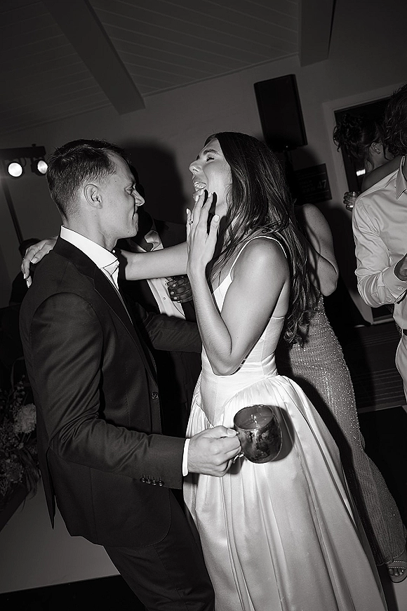 Wedding reception dancing as the bride and groom dancing, bride laughing in a satin dress while groom holds a drink amid party lights and guests