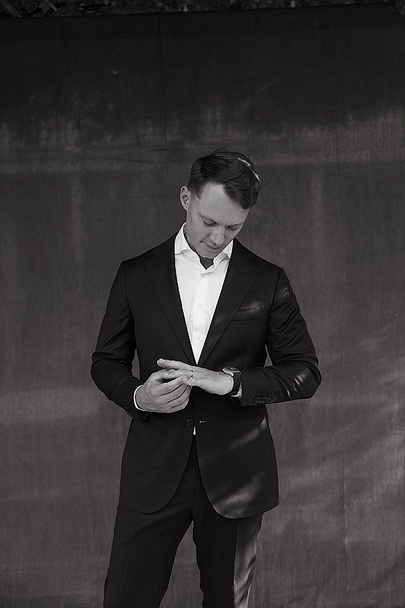 Groom portrait in a classic black suit adjusts his wedding band and checks a wristwatch against a dark textured wall backdrop