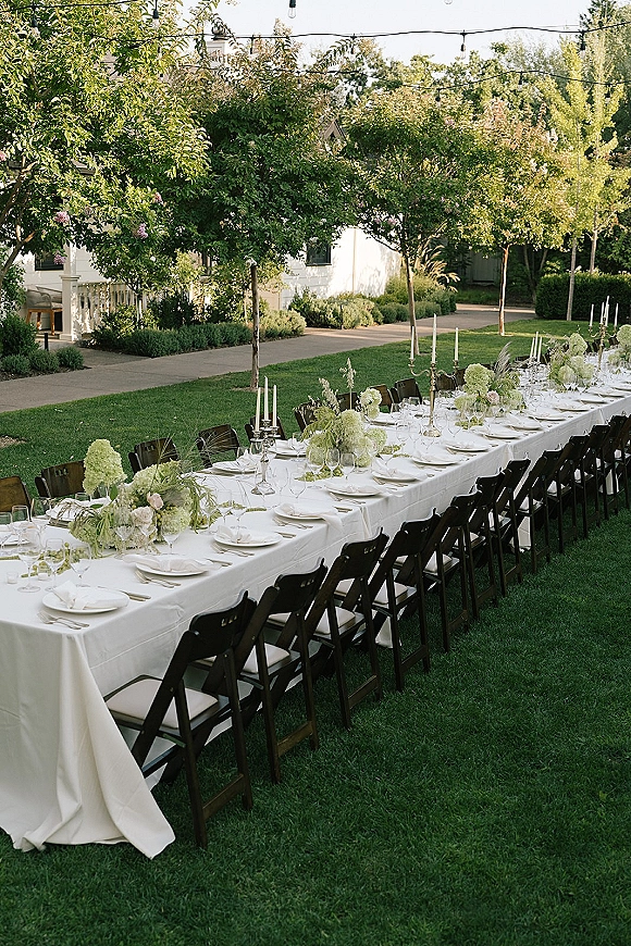 Reception tablescape with an outdoor wedding reception table set on a long banquet table, hydrangeas, greenery garland, taper candles, and string lights on a lawn near a white house