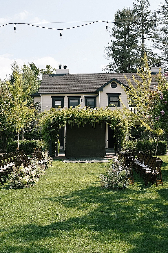 Ceremony setup for an outdoor wedding ceremony with wood folding chairs lining an aisle of florals beneath string lights on a grassy lawn