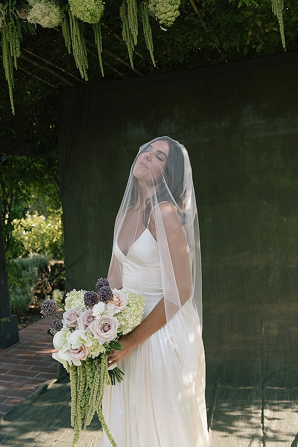 Bridal portrait of a bride with veil over face in a strapless satin wedding dress, holding a cascading rose and hydrangea bouquet in a garden setting