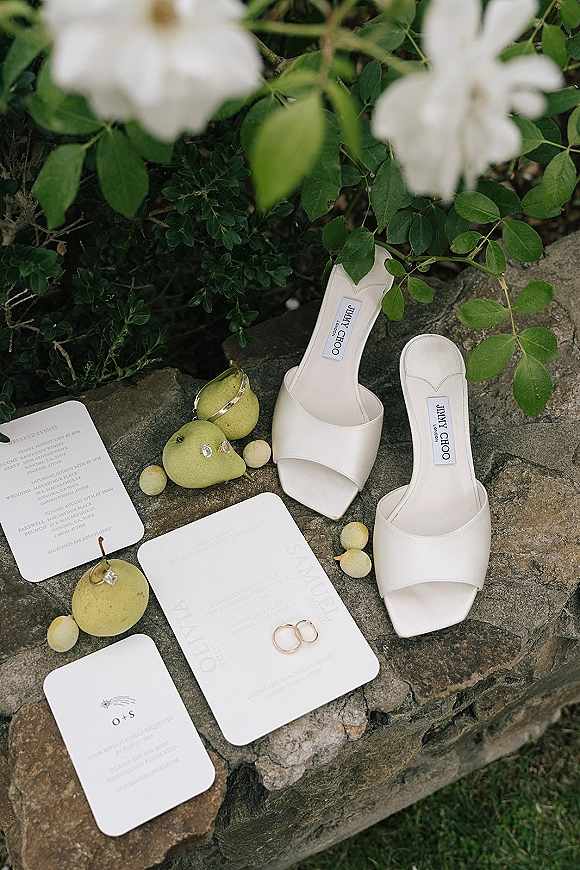Wedding flat lay with wedding invitation suite, white bridal sandals, rings, jewelry, pears and grapes on a stone ledge in garden foliage