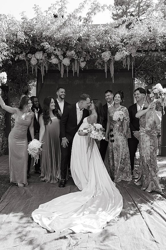 Wedding party photo of bride and groom with friends, bouquets and dress train, posing by a garden floral arch on a brick patio