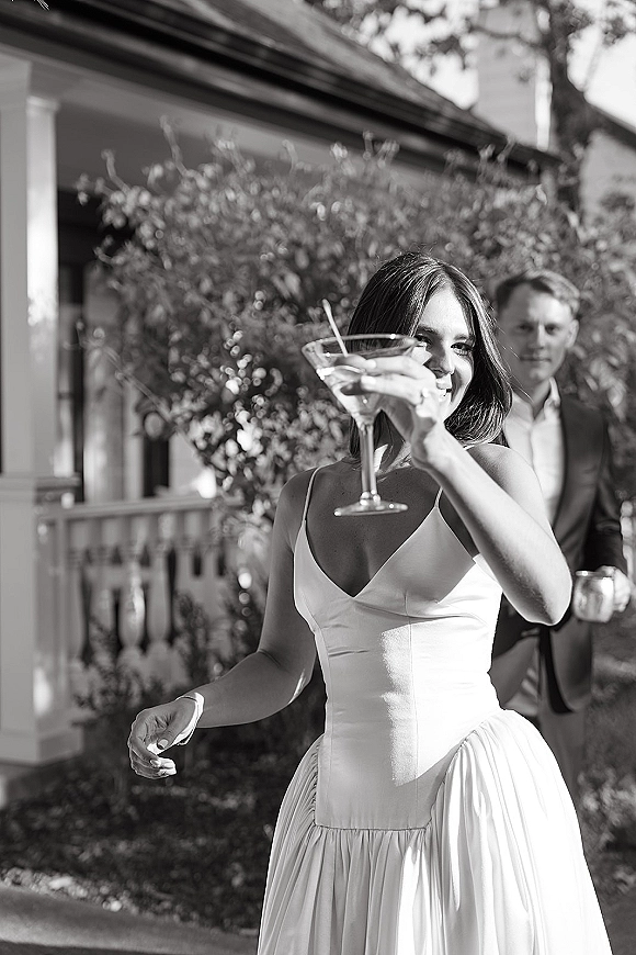 Wedding cocktail moment as bride in spaghetti-strap dress and groom in dark suit toast with martini on a porch by shrubs and railing
