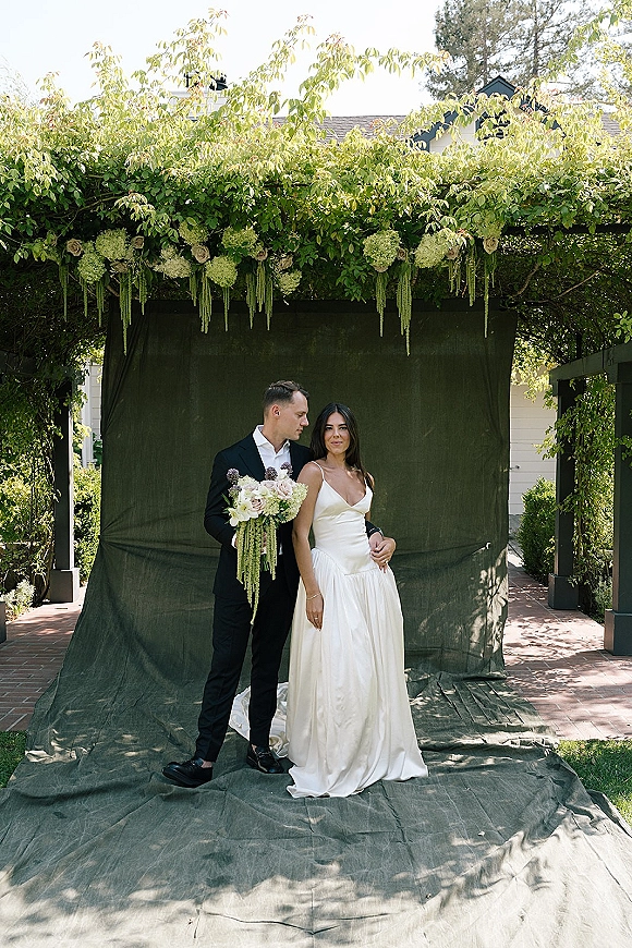 Couple portrait of bride in a satin gown and groom in a black suit holding a bouquet by a greenery-draped garden walkway backdrop