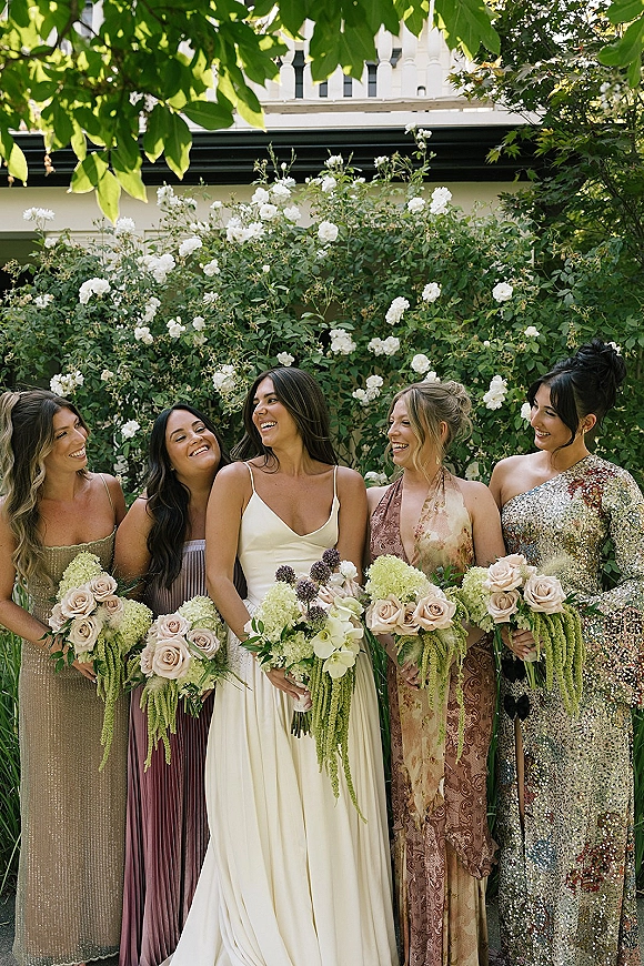 Bridesmaids portrait of women in mismatched dresses holding blush and white bouquets with cascading greenery in a white rose garden