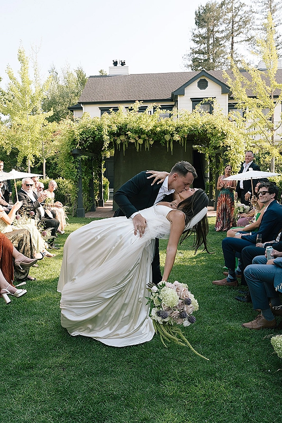 Wedding kiss as the groom dips the bride in her veil and wedding dress, bouquet in hand, on an outdoor aisle beneath a greenery arch
