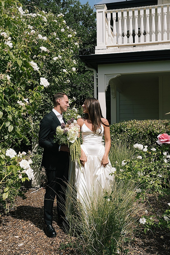 Couple portrait in a rose garden, bride in a satin gown holding a white-and-green bouquet beside groom in a black suit by a white porch