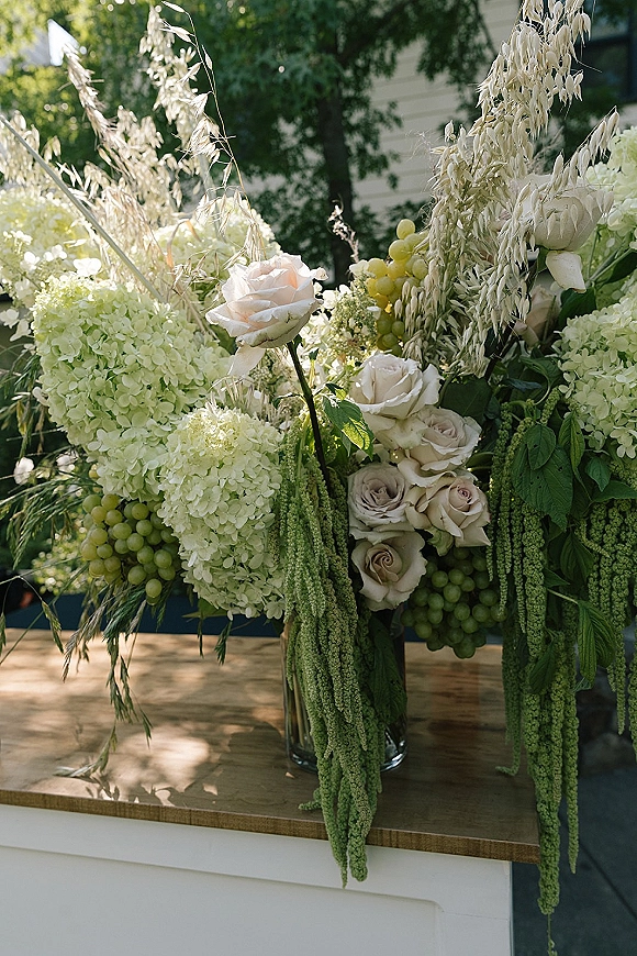 Wedding floral arrangement of white hydrangeas and roses with trailing amaranthus and greenery in a clear glass vase on a wooden table outdoors