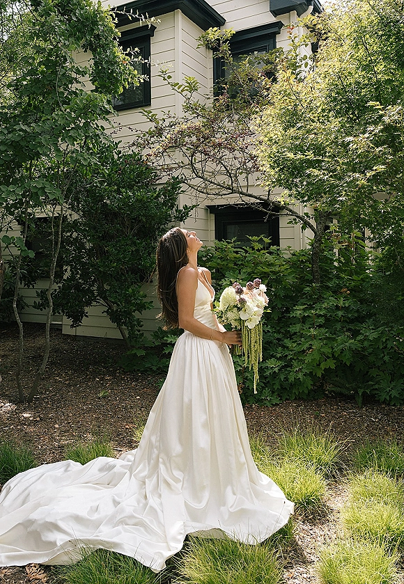 Bridal portrait of a bride holding bouquet, looking up in a strapless satin wedding dress with long train in a garden by a house exterior
