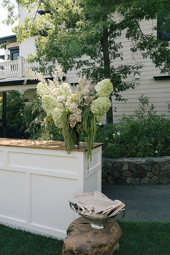 Wedding bar decor on a white bar setup with hydrangeas, roses, hanging greenery, and grapes on a lawn outside a house