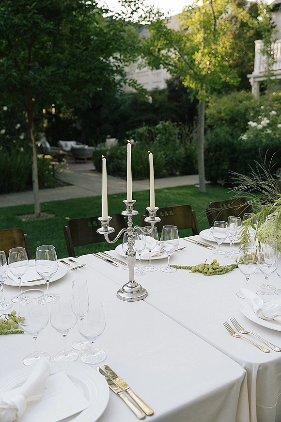 Reception tablescape with a white tablecloth, silver candelabra and taper candles, gold flatware, and grapes on a garden lawn patio setting