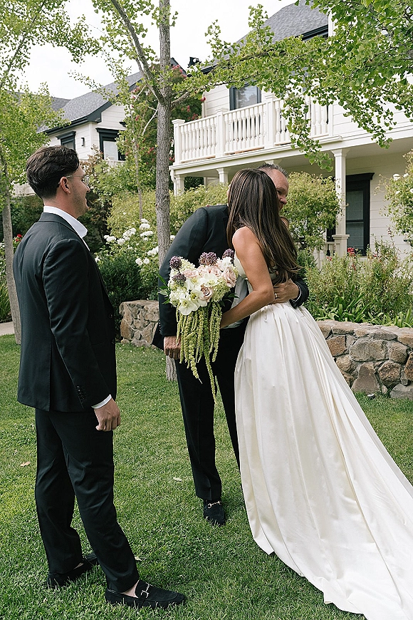 Wedding first look as the groom reaction turns into a hug, bride in a strapless dress holding a white and green bouquet on a garden lawn
